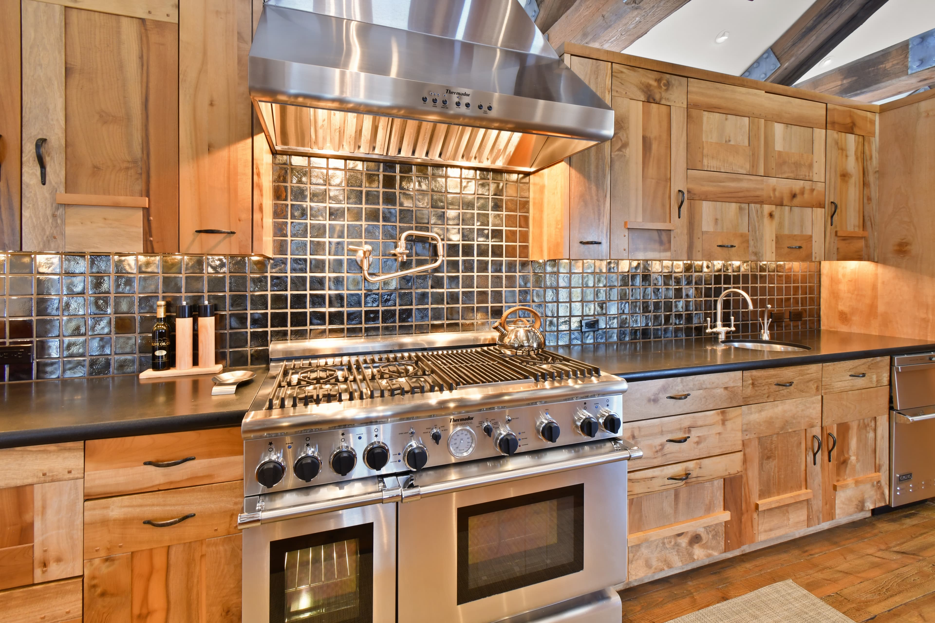 Black tile walls and wooden cabinetry surround a wide luxury oven range combo, with a industrial vent above.