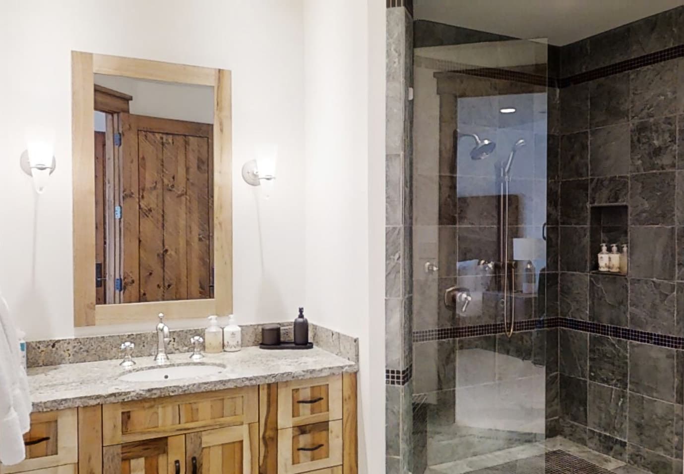 Black tiles highlight the walk-in shower of a bathroom, next to a simple sink and mirror.