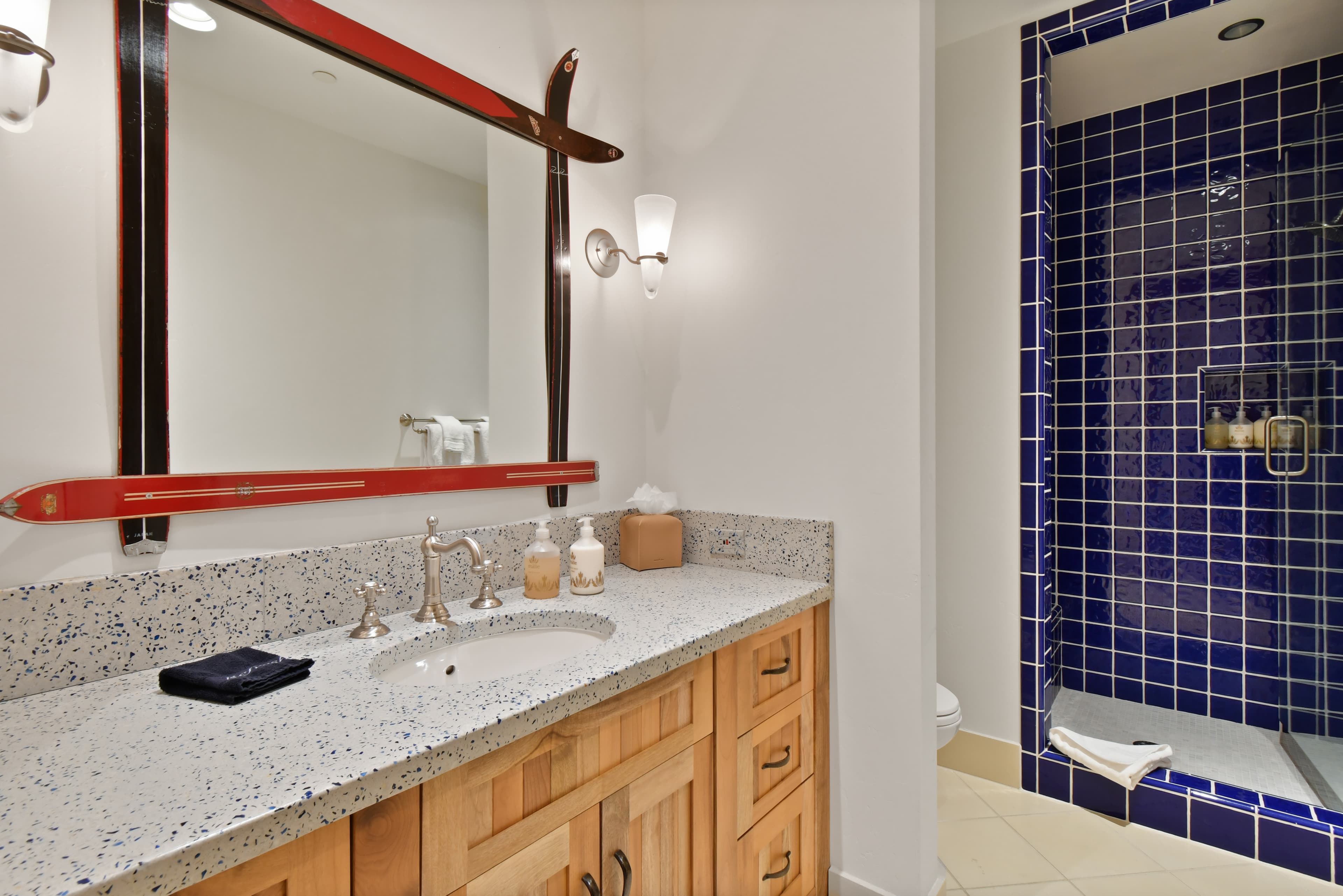 A mirror with a decorative frame sits above a simple sink counter, next to a blue-tile walk-in shower.